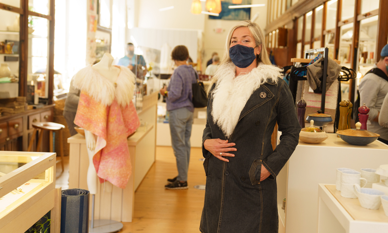 Woman in mask trying on coat at Maine Craft Portland, Photo Courtesy of Good Maine Woman in mask trying on coat at Maine Craft Portland, Photo Courtesy of Good Maine